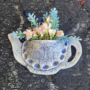 May include: A blue and white ceramic teapot-shaped vase with a floral design, filled with pink and blue artificial flowers. The vase has a handle and spout, and is decorated with a lace-like pattern.