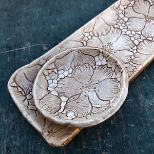 May include: A set of handmade ceramic serving dishes. The set includes a rectangular tray and a small round dish, both featuring a detailed floral pattern in shades of beige and brown. The dishes are placed on a dark teal surface.