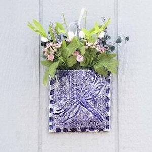 May include: Purple ceramic wall planter with a dragonfly design and a small bouquet of pink and white flowers.
