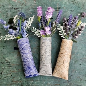 Puede incluir: Tres jarrones de cerámica con un patrón de encaje, cada uno con un ramo de flores moradas diferente. Los jarrones son de color azul, gris y beige.