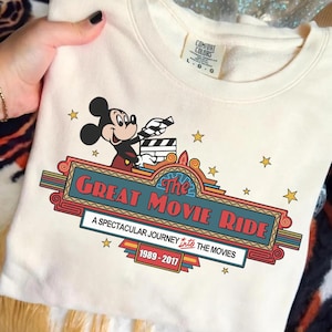 Puede incluir: Sudadera crema con Mickey Mouse sosteniendo una claqueta de cine. El dise&ntilde;o incluye el texto "The Great Movie Ride" en rojo y azul, con los a&ntilde;os 1989-2017. Estrellas doradas rodean el gr&aacute;fico.