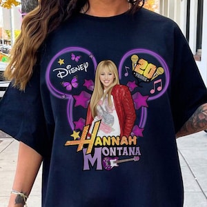 May include: Navy blue t-shirt featuring a graphic of Hannah Montana. The design includes the Disney logo, purple Mickey Mouse ears, stars, and the text "20 Years". The image also includes a picture of Hannah Montana wearing a red jacket.