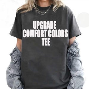 May include: Dark gray t-shirt with the text "UPGRADE COMFORT COLORS TEE" in white block letters. The shirt is worn with a light blue denim jacket.