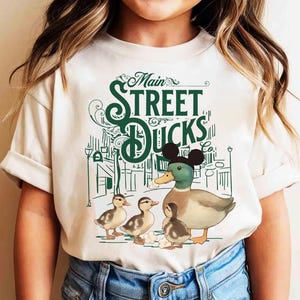 Op de afbeelding: Cr&egrave;mekleurig T-shirt met een groene afbeelding van een eendenfamilie en de tekst "Main Street Ducks Co.". De eend in het midden draagt een zwarte Mickey Mouse orenhoed. Het ontwerp bevat een straatbeeld met lantaarnpalen.
