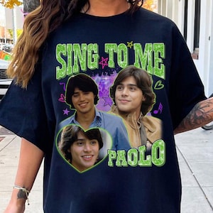 May include: A black t-shirt featuring a graphic print with the text SING TO ME and PAOLO in green glittery font. The design includes three photographic portraits of a person with dark hair, surrounded by purple and pink star and heart icons.
