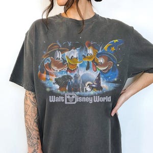 Może przedstawiać: Ciemnoszara koszulka z grafiką Goofy'ego, Kaczora Donalda i Myszki Miki przed zamkiem Kopciuszka. Tekst "Walt Disney World" jest wydrukowany pod obrazem.