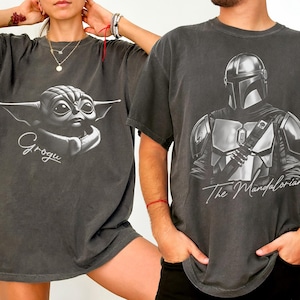 K&ouml;nnte beinhalten: Zwei anthrazitfarbene T-Shirts. Eines zeigt eine Grogu-Grafik mit dem Namen "Grogu" in Schriftform. Das andere zeigt ein Schwarz-Wei&szlig;-Bild von The Mandalorian mit dem Text "The Mandalorian".