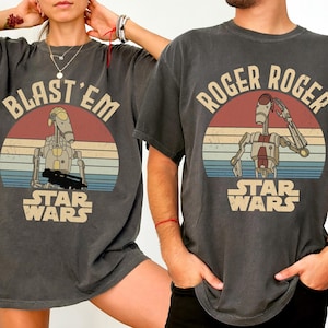 Puede incluir: Dos camisetas gris oscuro con gráficos de Star Wars. Una camiseta dice "BLAST'EM" con una ilustración de droide sobre un diseño de puesta de sol. La otra dice "ROGER ROGER" con un droide saludando sobre un diseño de puesta de sol. Ambas tienen el logo de Star Wars.