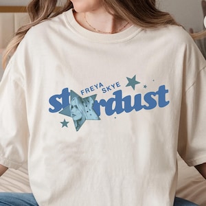 Puede incluir: Camiseta color marfil con la inscripción azul "Stardust" y un gráfico de estrella, junto con el nombre "Freya Skye". La camiseta tiene mangas cortas y un corte holgado.