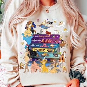 Puede incluir: Sudadera beige con una pila de libros coloridos con t&iacute;tulos y personajes de pel&iacute;culas de Disney. Los libros incluyen "La Bella y la Bestia", "Aladd&iacute;n", "101 D&aacute;lmatas", "Pocahontas" y "El Rey Le&oacute;n". Varios personajes de dibujos animados est&aacute;n representados.