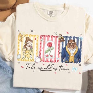 Puede incluir: Camiseta color crema con ilustraciones de Bella, una rosa en una campana de cristal y la Bestia. El diseño incluye el texto "Tale as old as time" en fuente cursiva, con estrellas y lazos decorativos.