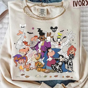 May include: An ivory crewneck sweatshirt featuring a graphic print of various cartoon characters dressed in Halloween costumes, including mummies, a skeleton, and a witch. The text COMFORT COLORS IVORY is printed in the upper right corner of the image.