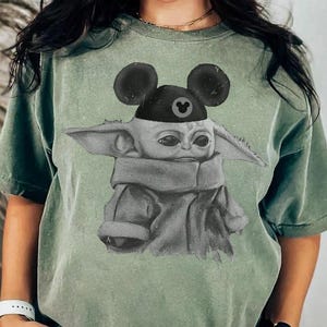 May include: A sage green short-sleeve t-shirt featuring a graphic print of a small, large-eared creature wearing a black cap with mouse ears. The creature is depicted in a sketch-like style with a gray color palette, wearing a draped garment.