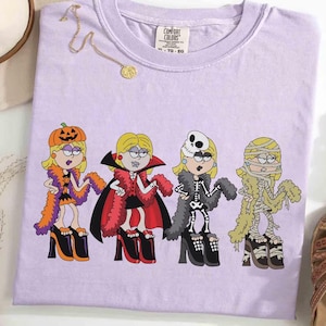 以下が含まれることがあります： ハロウィーンの衣装を着た4人の女性の漫画イラストが描かれた、薄紫色のComfort Colors Tシャツ。衣装には、カボチャ、吸血鬼、スケルトン、ミイラが含まれます。女性はハイヒールを履いています。