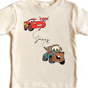 May include: Cream-colored t-shirt featuring cartoon images of Lightning McQueen and Mater from the movie Cars. The name "James" is written in cursive below the cars. The shirt has short sleeves.