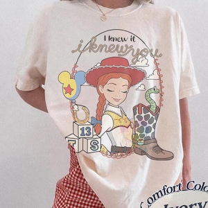 May include: An ivory t-shirt featuring a graphic of a cowgirl character with red hair, a red hat, and a yellow vest. The design includes the text I knew it i knew you, a balloon, a horseshoe, alphabet blocks with 13, and a patterned cowboy boot.