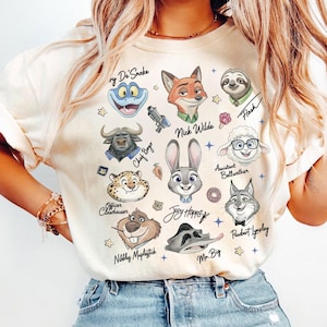 K&ouml;nnte beinhalten: Cremefarbenes T-Shirt mit Illustrationen von Zeichentrickfiguren aus Zootopia, darunter Nick Wilde, Judy Hopps und Flash. Das Shirt hat Charakternamen und Sternakzente. Das Shirt besteht aus einem weichen Material.