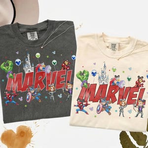 May include: Two t-shirts, one dark gray and one cream, featuring a Marvel design. The design includes Marvel characters, a castle, balloons, and the word "MARVEL" in red. Both shirts have a crew neck.