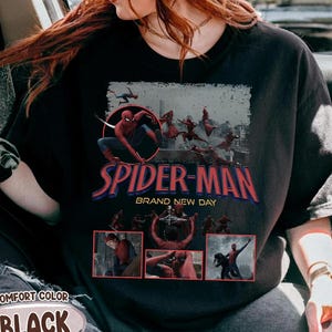 May include: Black t-shirt featuring a Spider-Man graphic. The design includes the words "SPIDER-MAN BRAND NEW DAY" in red and blue, with images of Spider-Man in action. The shirt also has the words "COMFORT COLOR BLACK" printed on it.