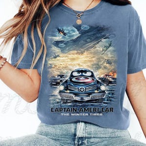 Puede incluir: Camiseta azul con un gráfico de un coche de dibujos animados con un escudo del Capitán América y el texto "CAPTAIN AMERI-CAR THE WINTER TIRES". El diseño incluye un avión y otros coches en el fondo.