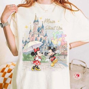以下が含まれることがあります： アイボリー色のTシャツ。お城を背景に、ミッキーとミニーマウスのカラフルなイラストが描かれています。ミニーマウスは傘を持ち、ミッキーマウスは風船を持っています。「Main Strat Usa」の文字がお城の上にあります。