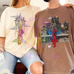 May include: Ivory and espresso-colored t-shirts. The ivory shirt features a cartoon of a tower with a princess hanging from her hair. The espresso shirt has a Spider-Man graphic with a city skyline background.