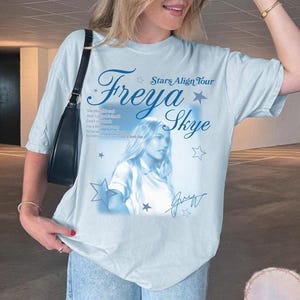 May include: Light blue t-shirt featuring the text "Freya Skye Stars Align Tour" in a stylized font, along with a portrait of a woman and her signature. The shirt has a relaxed fit and is made of a soft material.