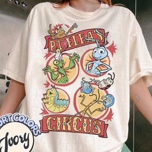 May include: Ivory-colored t-shirt with a colorful graphic featuring cartoon insects and the text "P.T. Flea's Circus." The design includes a red banner and circular frames. The shirt is a Comfort Colors brand.