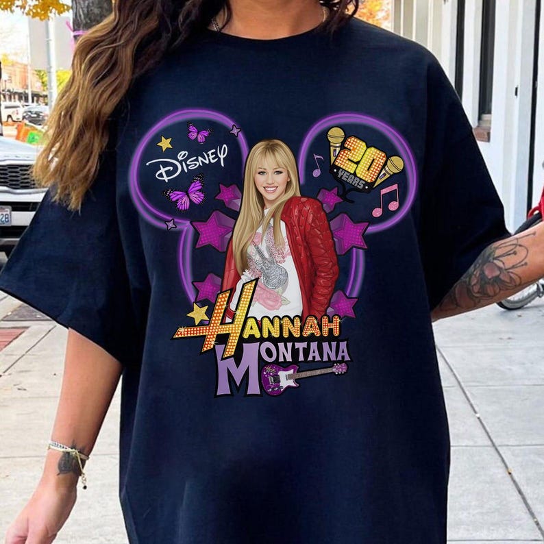 May include: Navy blue t-shirt featuring a graphic of Hannah Montana. The design includes the Disney logo, purple Mickey Mouse ears, stars, and the text "20 Years". The image also includes a picture of Hannah Montana wearing a red jacket.