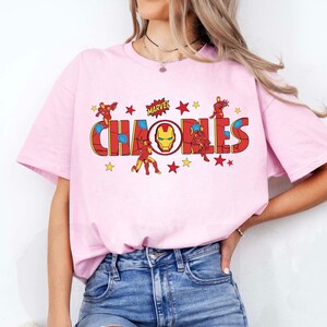 May include: A light pink t-shirt featuring the word "CHAOS" in a red and yellow comic book style font, with the Marvel logo above. The design includes illustrations of Iron Man and yellow stars.