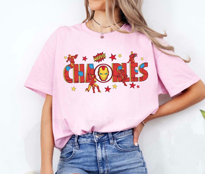 May include: A light pink t-shirt featuring the word "CHAOS" in a red and yellow comic book style font, with the Marvel logo above. The design includes illustrations of Iron Man and yellow stars.