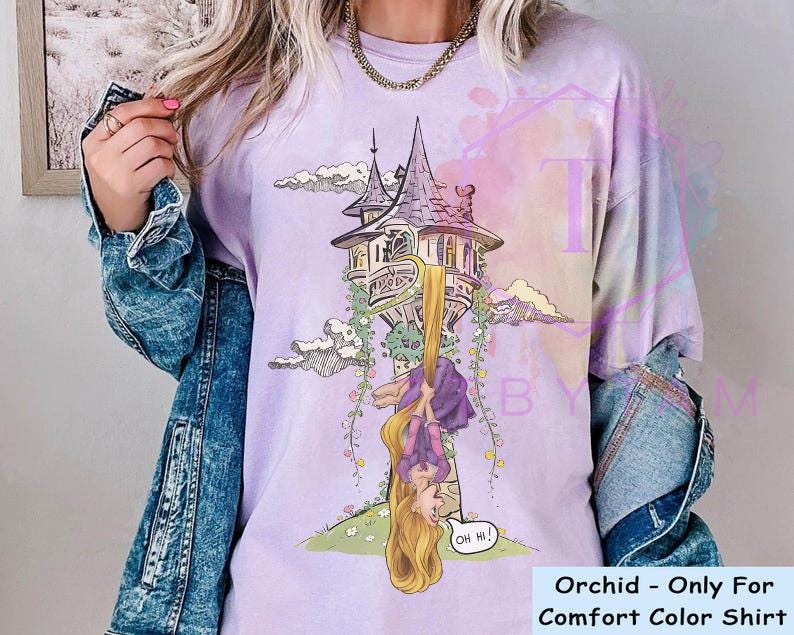 May include: Orchid-colored Comfort Color shirt featuring a cartoon illustration of a tower with a long-haired character hanging upside down. The character is wearing a purple dress and has long blonde hair. The shirt has the text "Oh Hi!" in a speech bubble.
