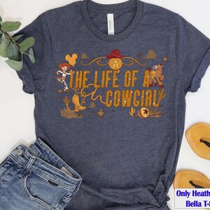 May include: Heather navy Bella T-shirt with the text "THE LIFE OF A COWGIRL" in gold glitter lettering. The design includes a cowgirl, a horse, a cowboy hat, and other western-themed graphics.