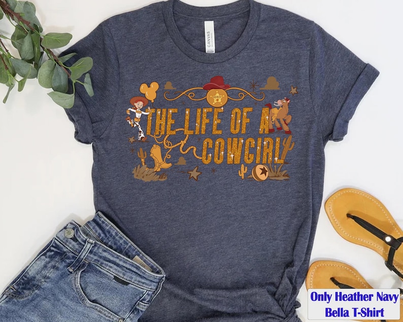 May include: Heather navy Bella T-shirt with the text "THE LIFE OF A COWGIRL" in gold glitter lettering. The design includes a cowgirl, a horse, a cowboy hat, and other western-themed graphics.