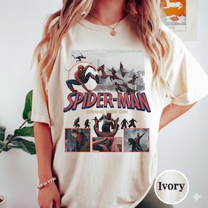 May include: Ivory-colored t-shirt featuring a Spider-Man graphic. The design includes the words "SPIDER-MAN BRAND NEW DAY" in red and blue, with multiple images of Spider-Man in action. The shirt is a casual wear item.