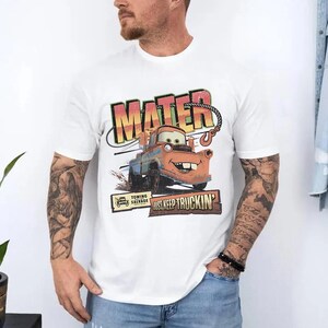 K&ouml;nnte beinhalten: Wei&szlig;es T-Shirt mit einem Aufdruck von Hook aus dem Film Cars. Das Design enth&auml;lt den Namen "MATER", das Bild von Hook und den Satz "JUST KEEP TRUCKIN'". Das T-Shirt hat kurze &Auml;rmel.