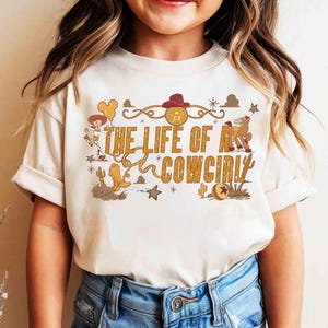 May include: Cream-colored t-shirt featuring the text "THE LIFE OF A COWGIRL" in a gold, Western-style font. The design includes a cowboy hat, boots, a horse, and other Western-themed graphics. The shirt is made of a soft material.