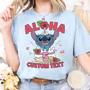 May include: Light blue t-shirt featuring a cartoon character holding up the word "ALOHA" above a cake with the number "5". The cake is decorated with flowers and the words "CUSTOM TEXT" are below the cake.