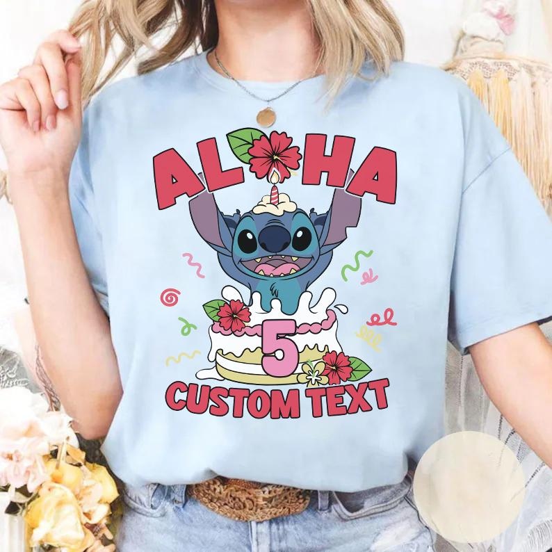 May include: Light blue t-shirt featuring a cartoon character holding up the word "ALOHA" above a cake with the number "5". The cake is decorated with flowers and the words "CUSTOM TEXT" are below the cake.