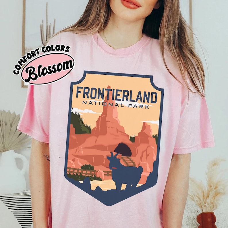 Puede incluir: Camiseta rosa claro con un dise&ntilde;o en forma de escudo con el texto "FRONTIERLAND NATIONAL PARK". El dise&ntilde;o incluye una cabra, monta&ntilde;as y un puente. La camiseta tambi&eacute;n tiene el texto "COMFORT COLORS Blossom".