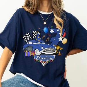 May include: Navy blue t-shirt featuring a cartoon car with a birthday hat, checkered flags, and the name "Patricia." The design includes a number 7, balloons, and confetti, creating a festive theme. The shirt is a casual wear item.