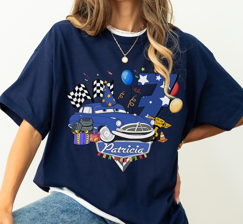 May include: Navy blue t-shirt featuring a cartoon car with a birthday hat, checkered flags, and the name "Patricia." The design includes a number 7, balloons, and confetti, creating a festive theme. The shirt is a casual wear item.