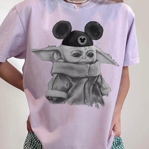 May include: A light purple t-shirt featuring a black and white graphic illustration of a small creature with large ears wearing a black hat with round mouse-like ears and a small circular icon in the center of the hat brim.
