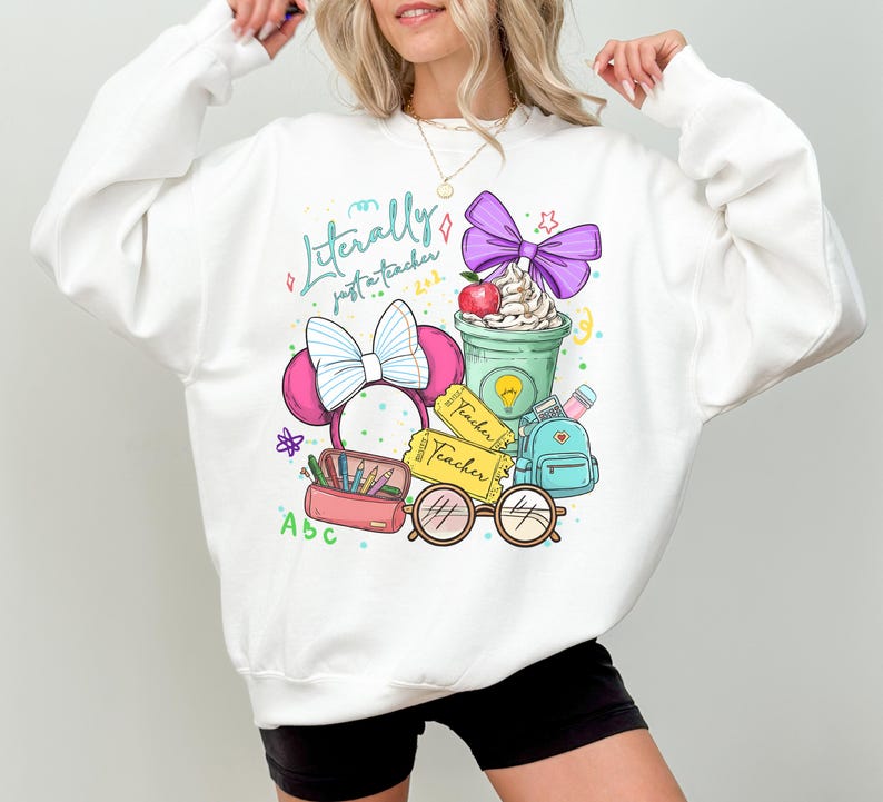 May include: White sweatshirt with a colorful graphic that reads "Literally just a teacher 2023." The graphic includes a coffee cup, apple, bow, glasses, pencil case, and backpack. The sweatshirt is a crew neck.