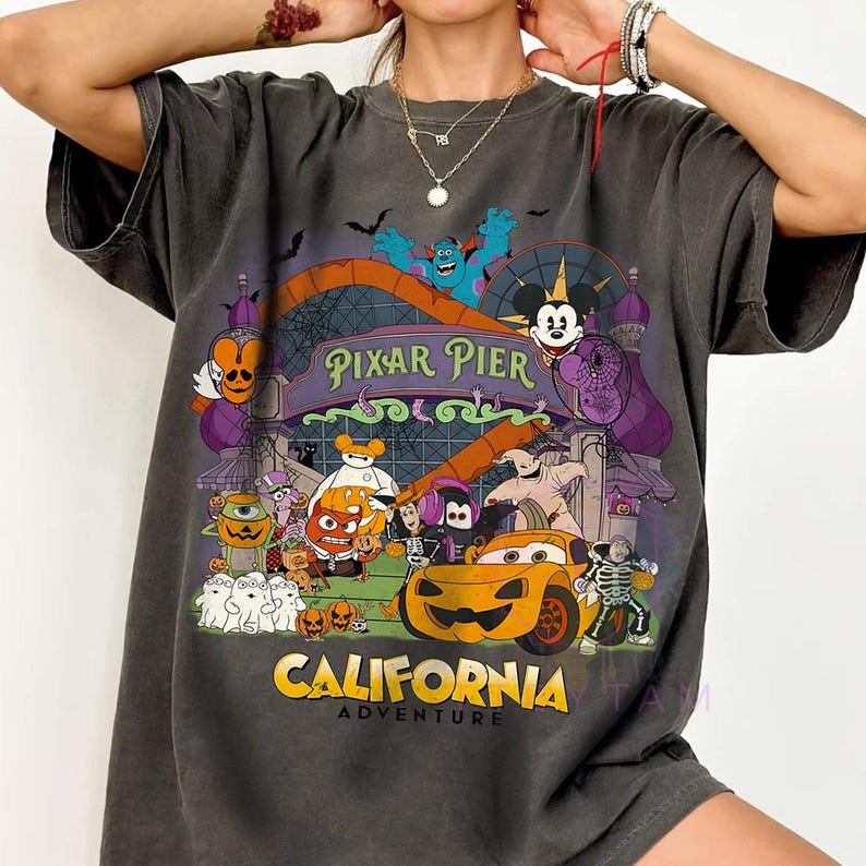 K&ouml;nnte beinhalten: Ein anthrazitfarbenes Kurzarm-T-Shirt mit einem farbenfrohen Grafikdruck von Pixar Pier im California Adventure, das verschiedene Zeichentrickfiguren in Halloween-Kost&uuml;men, K&uuml;rbisse, Geister und den Schriftzug Pixar Pier California Adventure zeigt.