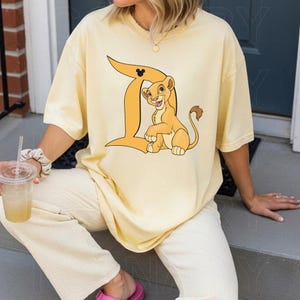 Peut inclure: Un t-shirt &agrave; manches courtes jaune p&acirc;le arborant une grande lettre D orange avec une petite ic&ocirc;ne noire de Mickey Mouse en haut. &Agrave; l'int&eacute;rieur de la lettre D se trouve une illustration graphique d'un jeune lionceau au pelage brun clair, au ventre cr&egrave;me et au bout de la queue brun fonc&eacute;.