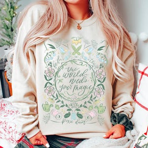 Pu&ograve; includere: Felpa beige con un design floreale e la scritta "the world needs your magic". Il design include fiori rosa e blu, foglie verdi e le parole "faith" e "trust". Sono incluse anche le parole "pixie dust".