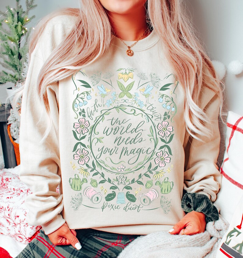 Pu&ograve; includere: Felpa beige con un design floreale e la scritta "the world needs your magic". Il design include fiori rosa e blu, foglie verdi e le parole "faith" e "trust". Sono incluse anche le parole "pixie dust".