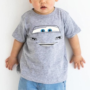 May include: A heather gray short-sleeve t-shirt featuring a graphic print of a cartoon car face. The design includes large white windshield eyes with blue pupils, black outlined headlights, and a wide, smiling mouth centered on the chest area.