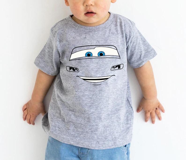 May include: A heather gray short-sleeve t-shirt featuring a graphic print of a cartoon car face. The design includes large white windshield eyes with blue pupils, black outlined headlights, and a wide, smiling mouth centered on the chest area.